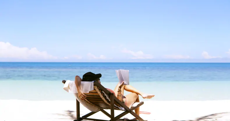 Pessoa relaxando em uma cadeira de praia, lendo um livro, com o oceano azul ao fundo.
