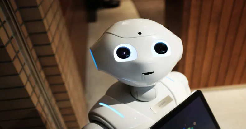 Robô branco com olhos grandes e pretos, detalhes azuis, segurando um tablet.