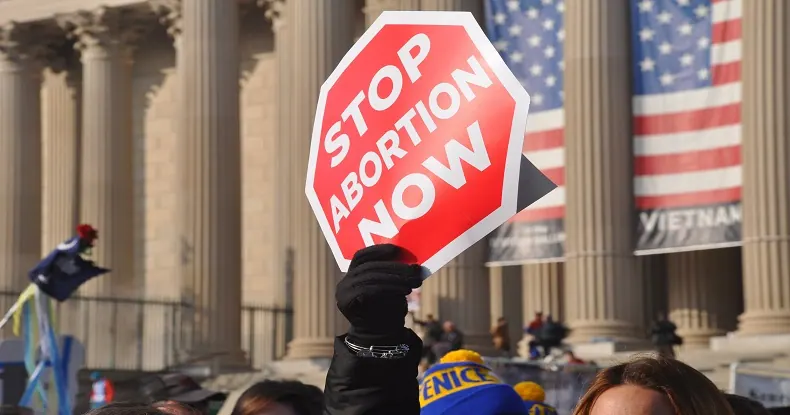 Pessoas em protesto, com uma mão levantando uma placa vermelha hexagonal escrito STOP ABORTION NOW.