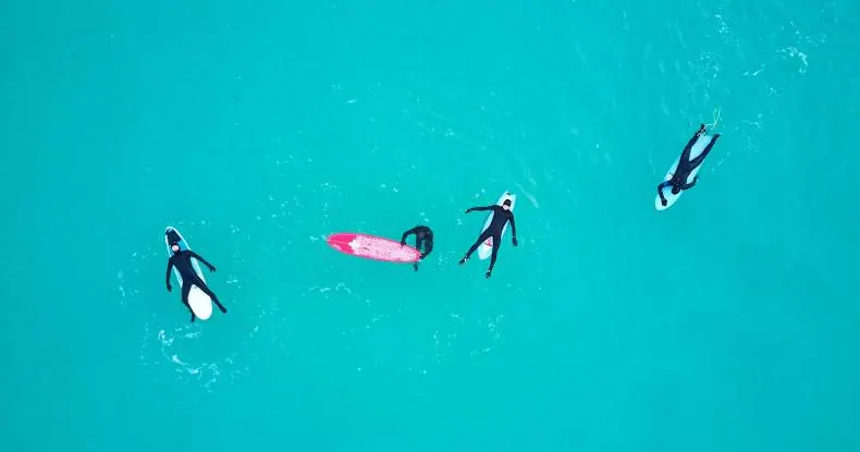 Quatro surfistas em roupas de mergulho flutuando em águas azul-turquesa, vista aérea.