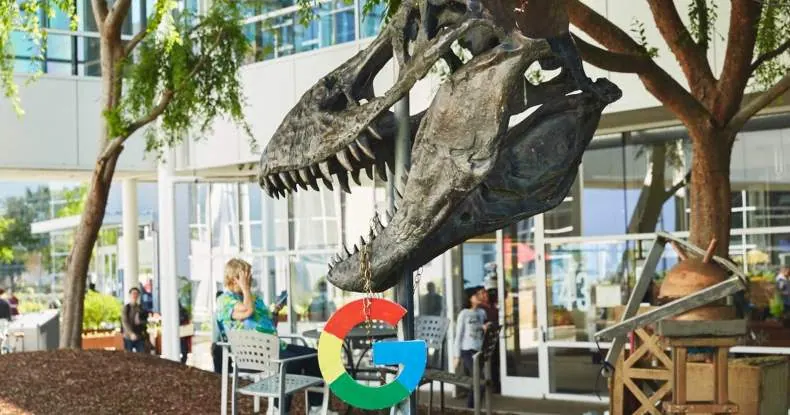 Esqueleto de dinossauro com o logotipo do Google pendurado na boca, pessoas ao fundo.