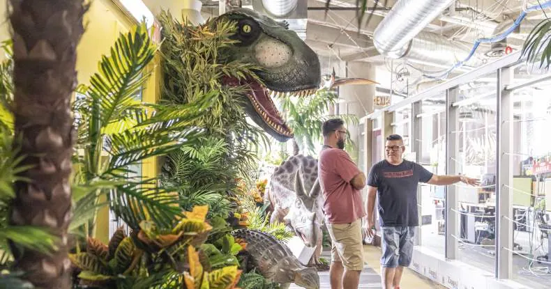 Dois homens conversam em um corredor com decoração de dinossauros e plantas.