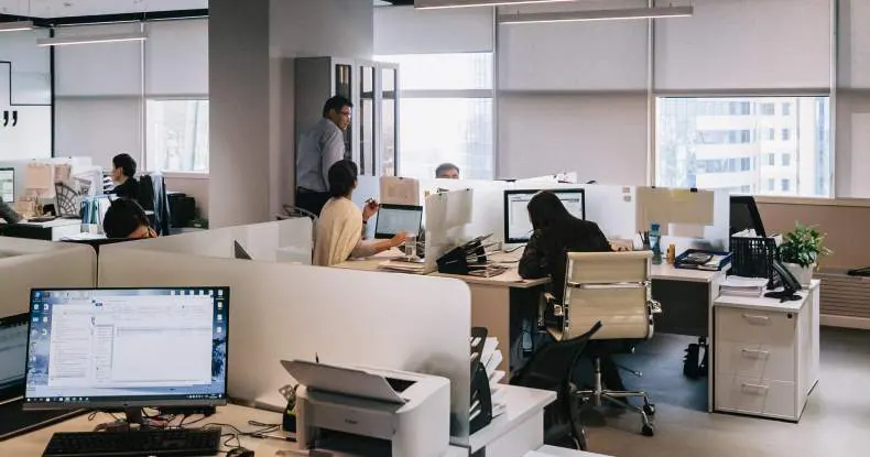 Pessoas trabalhando em um escritório moderno com cubículos, computadores e janelas grandes.