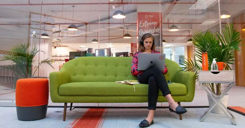 Mulher jovem com fones de ouvido trabalhando em um laptop no sofá verde em um escritório moderno.