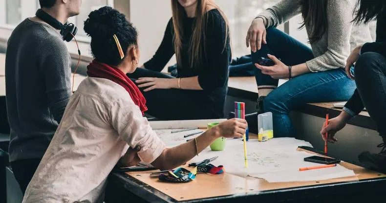 Pessoas colaborando em uma mesa, com canetas coloridas e papéis espalhados.