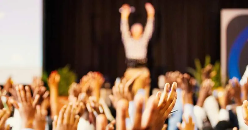 Pessoas com as mãos levantadas em um evento, com um palestrante ao fundo.