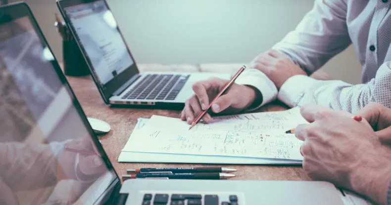 Duas pessoas trabalhando em uma mesa com laptops e papéis, escrevendo e apontando.