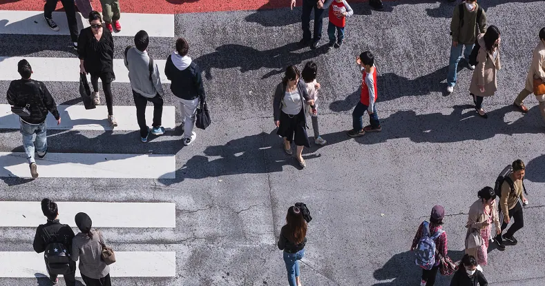 Pessoas diversas caminhando em uma faixa de pedestres e na rua, vista de cima.