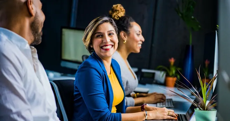Três colegas de trabalho sorrindo e usando computadores em um escritório moderno.