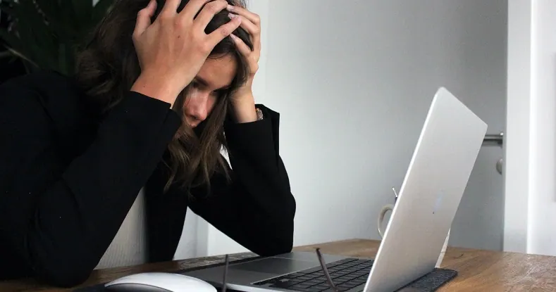 Mulher com as mãos na cabeça, olhando para baixo, sentada em uma mesa com um laptop.