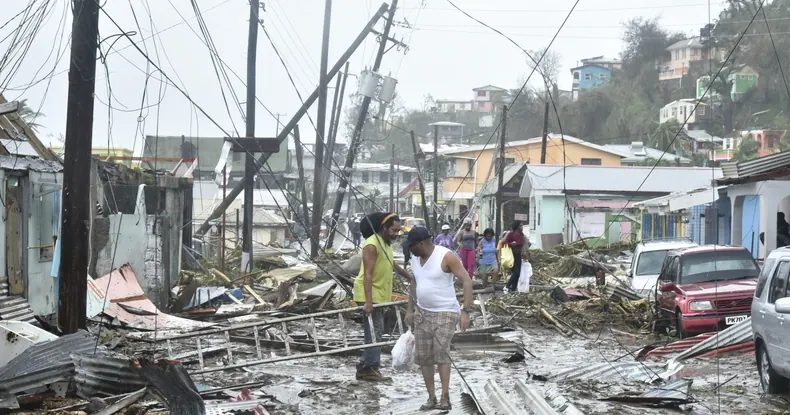 Pessoas andam por uma rua devastada, com postes caídos e casas destruídas.