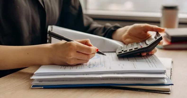 Mãos de uma pessoa escrevendo em documentos com uma calculadora ao lado.