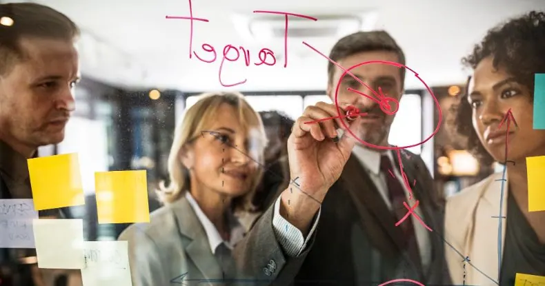 Quatro colegas de trabalho colaboram em um quadro de vidro com anotações e gráficos.