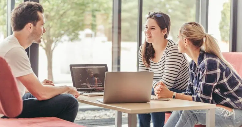Três colegas de trabalho conversando em uma mesa com dois notebooks em um escritório moderno.