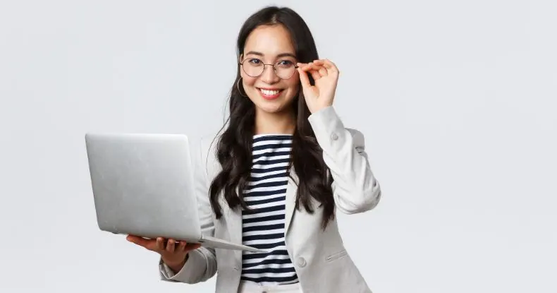 Mulher asiática sorridente, de óculos e blazer, segurando um notebook prateado.