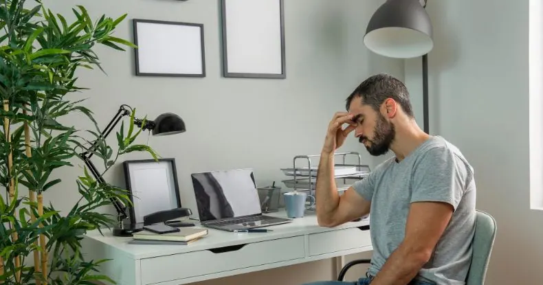 Homem barbudo, com expressão de cansaço, sentado em uma mesa de escritório com laptop.