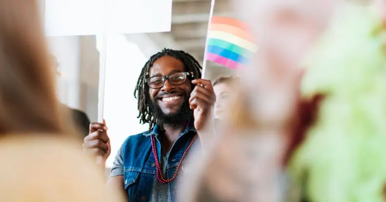 Homem negro sorridente com dreadlocks e óculos, segurando uma bandeira do arco-íris e um cartaz em um evento.
