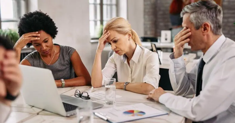 Três colegas de trabalho, visivelmente estressados, sentados em uma mesa de reunião.