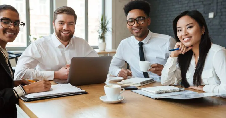 Quatro colegas de trabalho sorridentes sentados em uma mesa com laptops e xícaras de café.