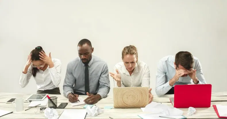 Quatro colegas de trabalho estressados e sobrecarregados em suas mesas com papéis amassados.