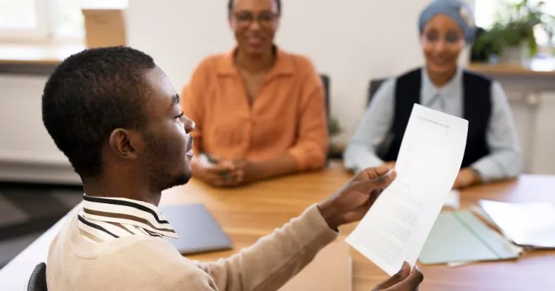 Homem negro sorrindo, segurando um documento, com duas mulheres ao fundo.