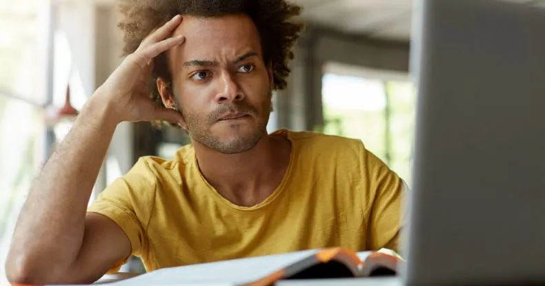 Homem com cabelo encaracolado e camisa amarela olhando para a tela do laptop com expressão pensativa.