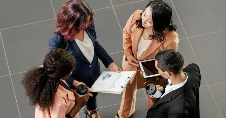 Quatro pessoas de negócios em pé, olhando para gráficos em uma prancheta.