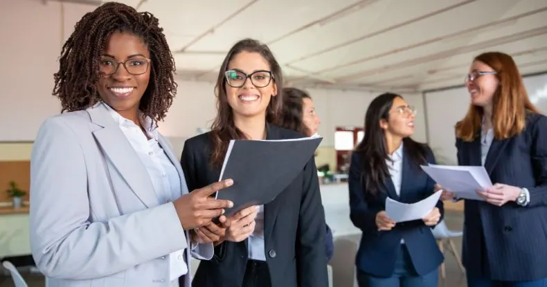 Cinco mulheres sorridentes em ternos de negócios segurando documentos em um escritório.
