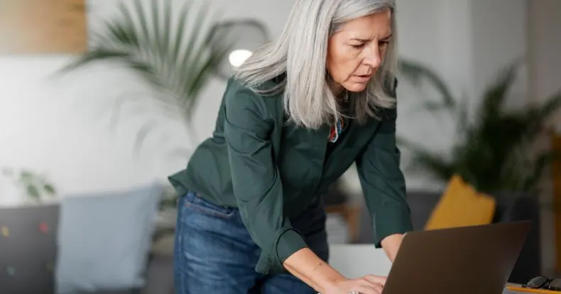 Mulher idosa de cabelos grisalhos e camisa verde escura olhando para um laptop.