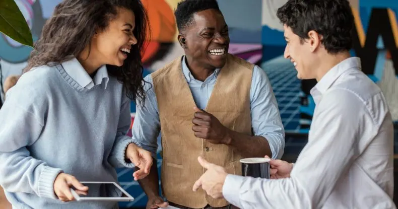 Três colegas de trabalho sorrindo e conversando em um escritório colorido.