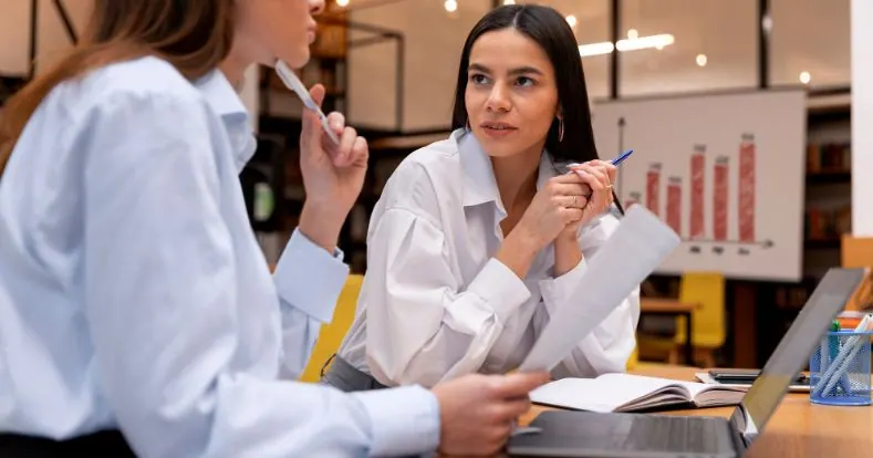 Duas mulheres em camisas brancas discutem documentos e um laptop no escritório.