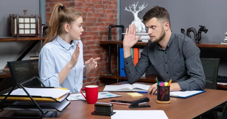 Dois colegas de trabalho discutindo em uma mesa de escritório, com o homem levantando a mão.