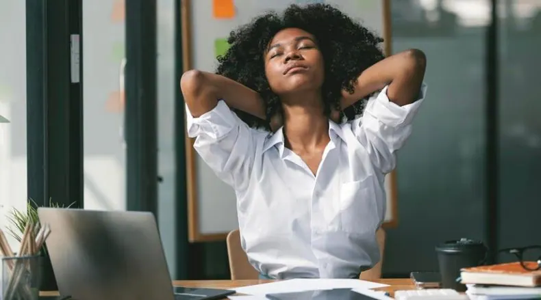 Mulher jovem de pele escura e cabelo cacheado alongando-se em uma mesa de escritório.