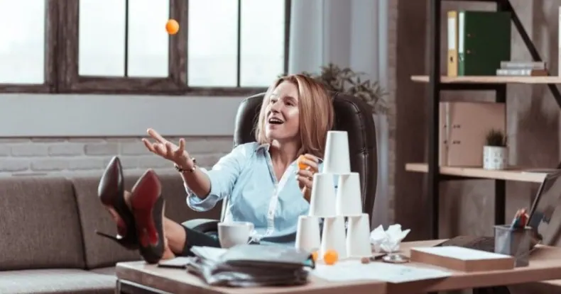 Mulher sorridente jogando bolinhas de pingue-pongue no escritório com os pés na mesa.