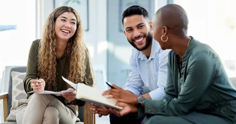 Três colegas de trabalho sorrindo e conversando em um ambiente de escritório descontraído.
