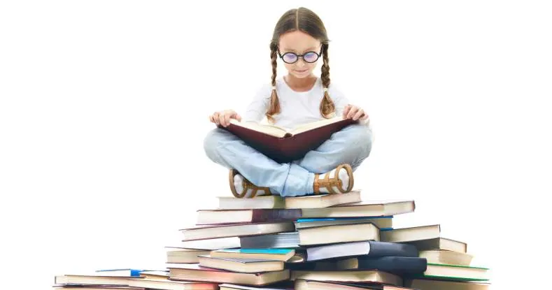 Menina de óculos sentada de pernas cruzadas lendo um livro sobre uma pilha de livros.