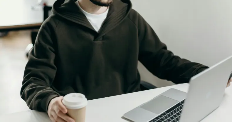 Homem de moletom escuro trabalhando em um laptop com uma xícara de café.