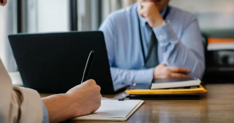 Duas pessoas em uma mesa, uma escrevendo e outra observando, com um laptop.