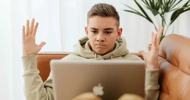 Adolescente de capuz cinza, com expressão de frustração, gesticula com as mãos enquanto olha para um laptop.