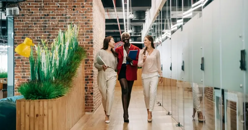 Três mulheres sorridentes caminhando por um corredor moderno de escritório.