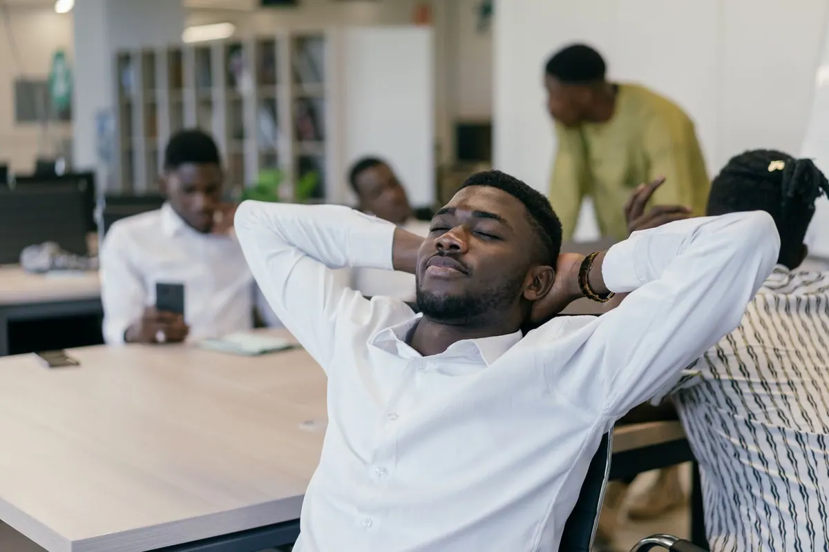 Homem negro de camisa branca relaxando em uma cadeira no escritório, olhos fechados.