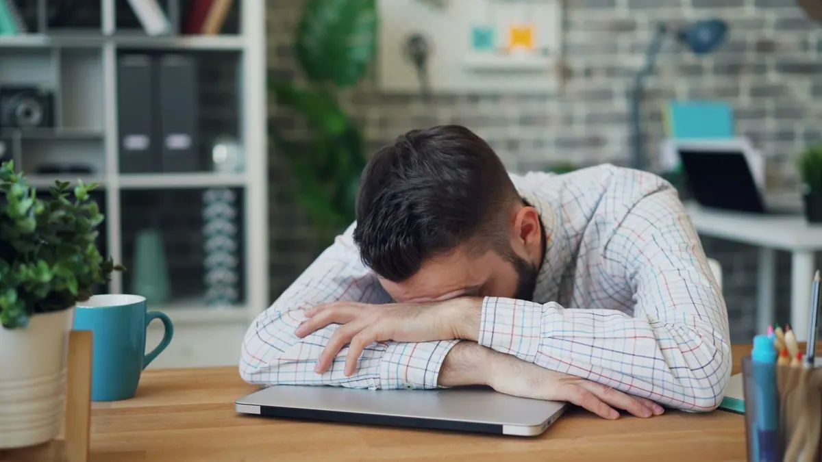 Homem com a cabeça deitada sobre o notebook na mesa do escritório.