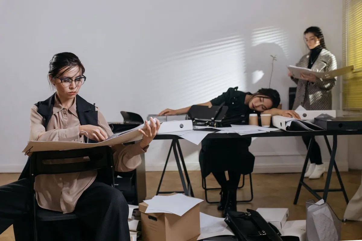 Mulheres exaustas e sobrecarregadas em um escritório desorganizado, simbolizando esgotamento.