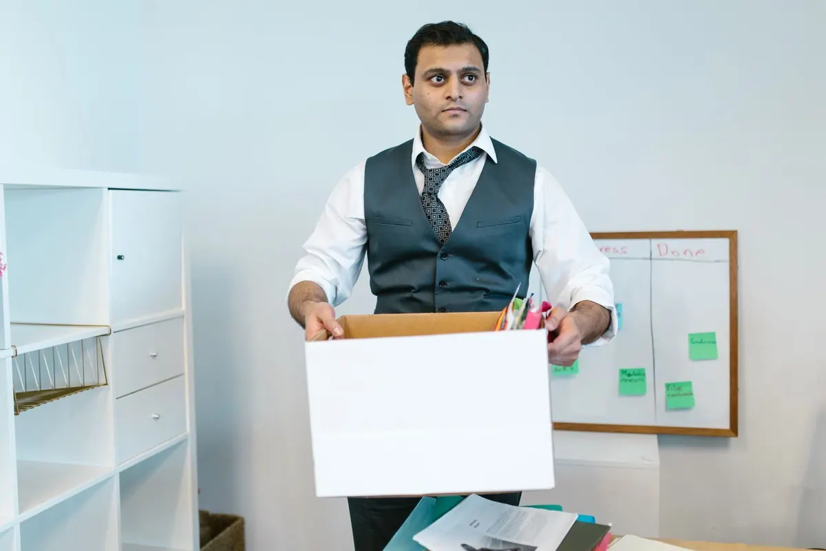 Homem de terno e gravata, com expressão séria, carregando uma caixa de pertences de escritório.