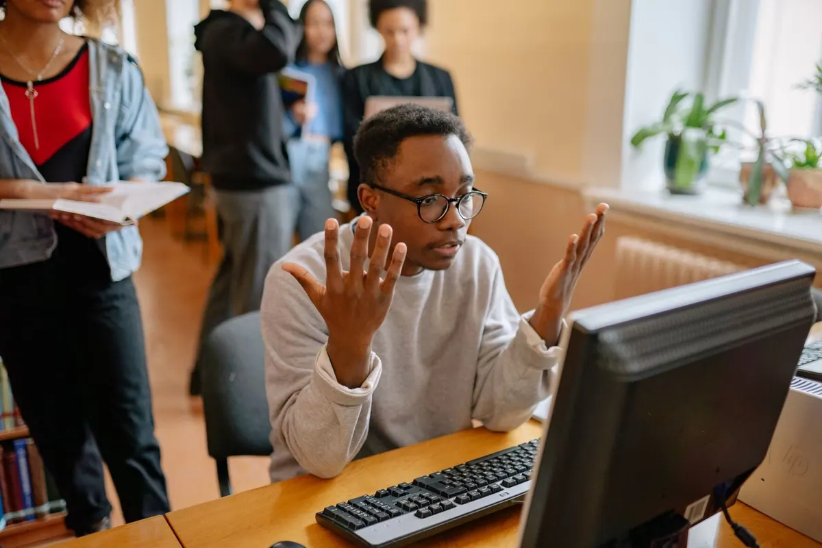 Homem negro confuso gesticula em frente ao computador, com colegas ao fundo.