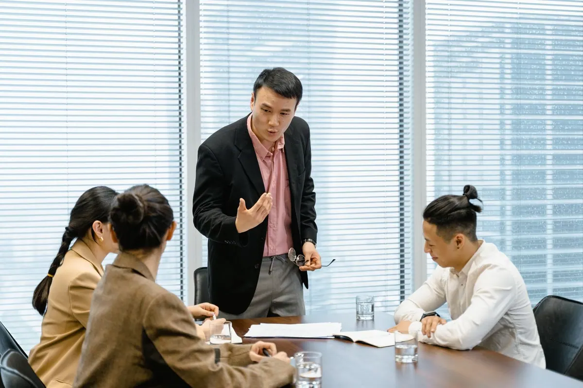 Líder asiático de pé, gesticulando, falando com três funcionários sentados.