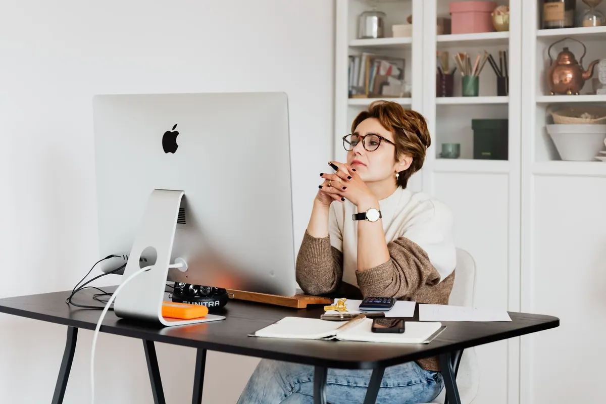 Mulher de óculos sentada à mesa do escritório, olhando pensativamente para a tela do computador.