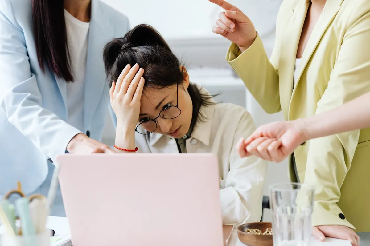 Mulher asiática estressada no trabalho, com colegas apontando para a tela do laptop.