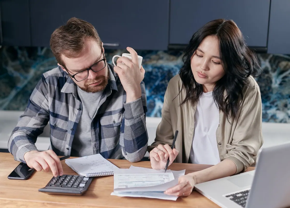 Casal calculando finanças domésticas com calculadora, papéis e laptop.