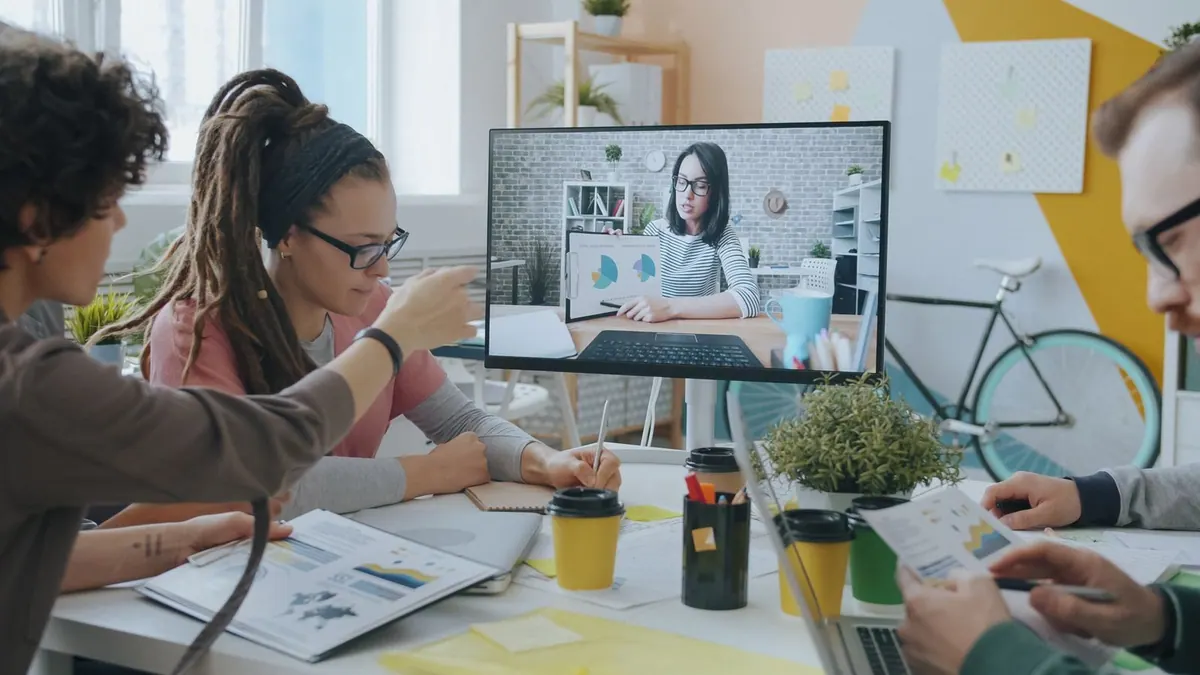 Pessoas em um escritório moderno colaborando em uma videochamada, com uma tela grande mostrando um colega.